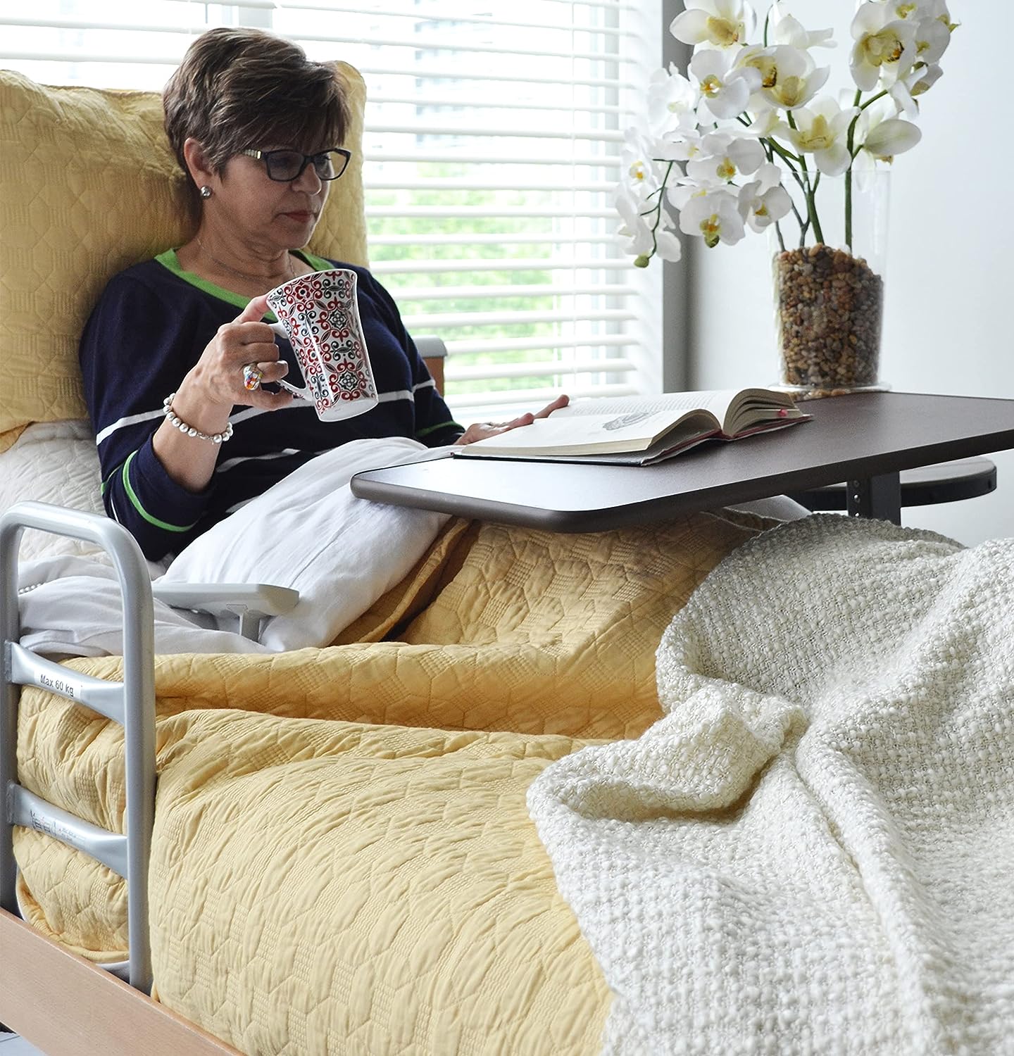 Lady lying in bed using Invacare's item number 6417, Hospital-Style, Brown Overbed Table reading a book and holding a mug.